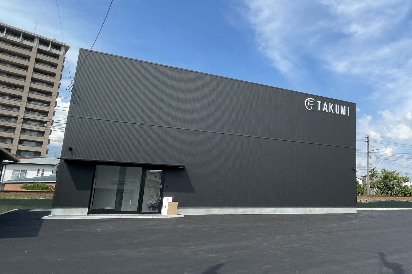松山市平井町にある株式会社匠の本社。黒い外壁の箱型の社屋で、壁に「TAKUMI」の看板がある。手前は駐車場